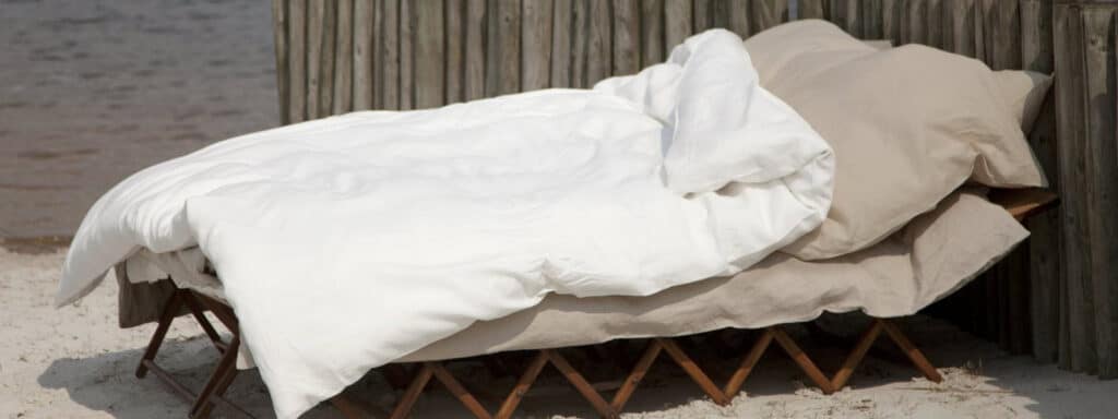 Ein gemütliches Outdoor-Bett mit beigen Laken und Kissen, bedeckt mit einer flauschigen weißen Decke, ruht auf einem klappbaren Holzrahmen in der Nähe eines rustikalen Holzzauns auf sandigem Boden - perfekt zum Entspannen und Staunen: Wie pflege ich Leinenbettwäsche richtig?.