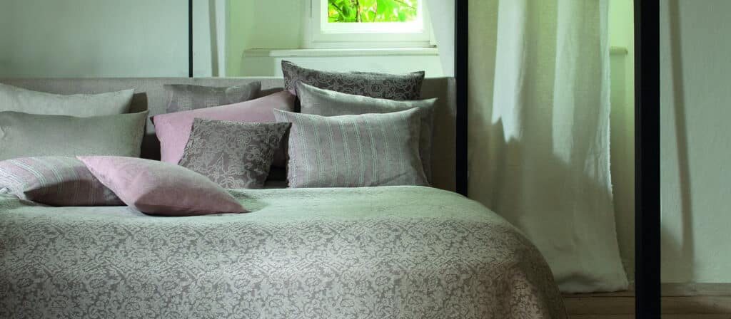 Ein ordentlich gemachtes Bett mit einer geometrischen Muster-Bettdecke und mehreren dekorativen Kissen in Grau- und Violetttönen, in der Nähe eines Fensters mit weißen Vorhängen, die das natürliche Licht hereinlassen.