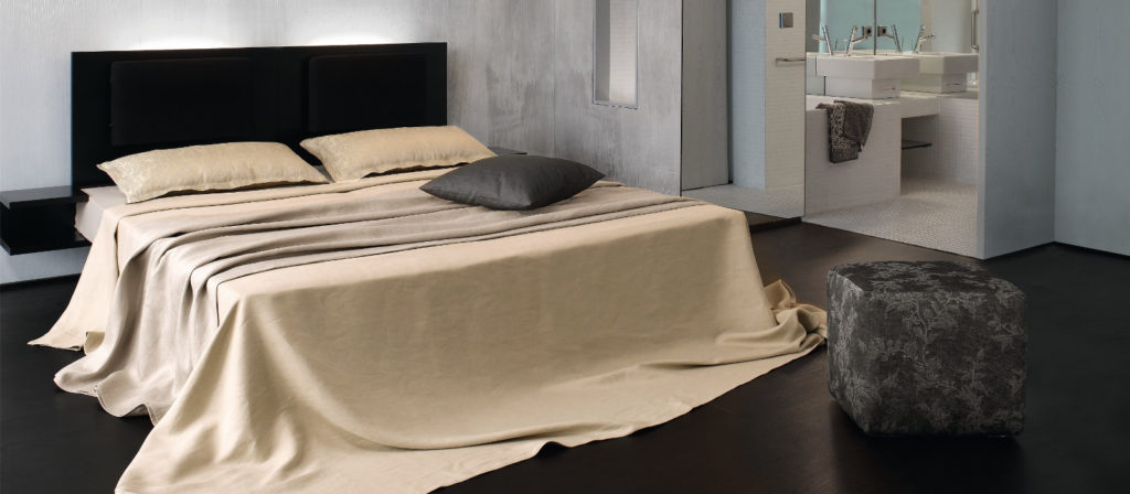 A modern bedroom with a large bed covered in plain beige fabric—just bedding, two pillows, and a gray cushion. Nearby is a gray textured stool, which offers a view of a simple bathroom with a white sink and mirror.