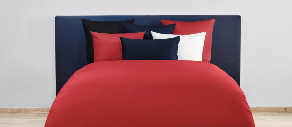 A neatly made bed with a red duvet, matching red and navy blue pillowcases, a white pillowcase, and a dark blue headboard, set against a light-colored wall—Uni Stoffe provides style and comfort, exclusively for your bedroom.