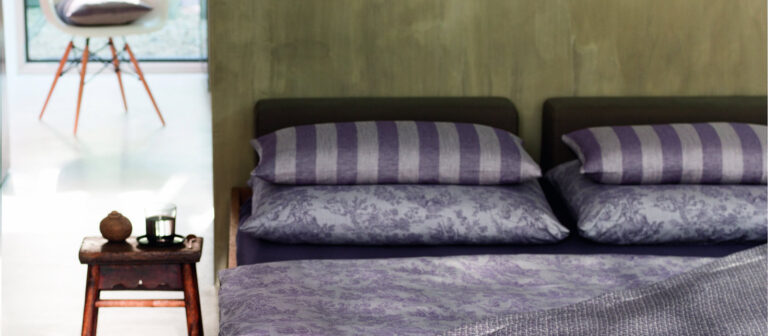 Ein ordentlich gemachtes Bett mit lila und grau gestreiften Kissen - der Streifen zeigt einen eleganten Kontrast -, gepaart mit einer gemusterten lila Tagesdecke, neben einem kleinen hölzernen Beistelltisch mit einer Kerze in einem modernen, sanft beleuchteten Schlafzimmer.