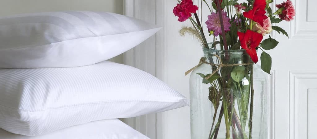 Two white linen weave cushions lie next to a clear glass vase filled with colourful flowers in front of a white wall and door.
