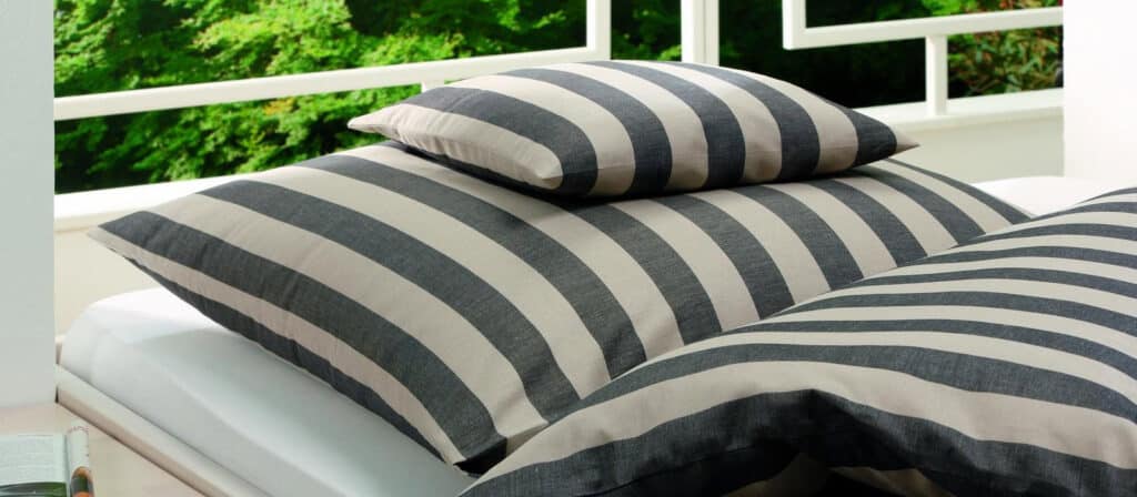Black and gray striped pillows and a matching blanket lie on a white bed outdoors; the stripes create elegant contrasts, while green trees can be seen in the background behind a white railing.