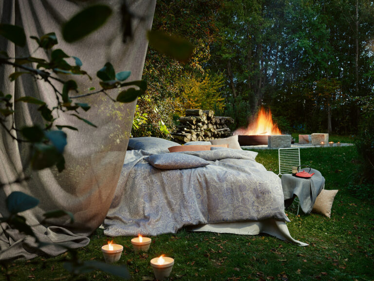 A cozy outdoor bed with patterned bedding, surrounded by candles, sits on the grass next to a fire pit, with stacked firewood and trees in the background creating a peaceful, inviting nighttime atmosphere.