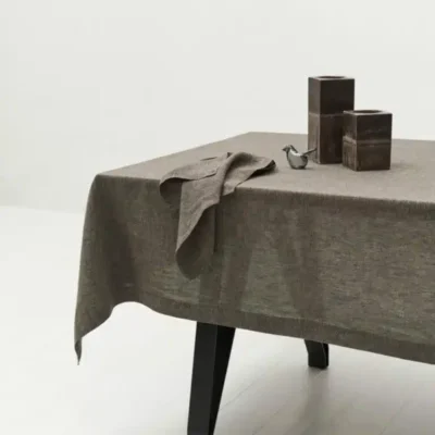 Ein mit einem braunen Tischtuch bedeckter Tisch mit einer gefalteten Serviette, zwei quadratischen dunkelbraunen Vasen und einem kleinen Metallvogel als Ornament auf dem Tisch. Der Tisch steht auf schwarzen Beinen in einem minimalistischen, weißen Raum.