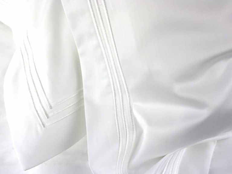 Nahaufnahme eines weißen Bettlakens mit einem dezenten, abgesteppten Bordürenmuster, das die weiche Struktur des Stoffes und die glatten Falten zeigt.