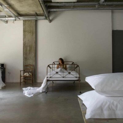 Eine Frau sitzt auf einem Metallbett in einem minimalistischen Raum im Industriestil mit Betonboden und freiliegenden Rohren. Große weiße Kissen sind im Vordergrund gestapelt. Von links strömt Licht herein.
