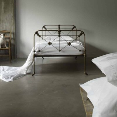 Ein metallgerahmtes Bett mit weißer Bettwäsche steht in einem minimalistischen Raum mit Betonboden. Neben dem Bett steht ein Stuhl mit einem kleinen Hund darauf, und weiße Laken ziehen sich vom Bett bis zum Boden. Zwei Kissen befinden sich im Vordergrund.