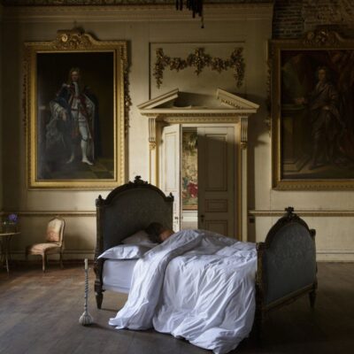 Eine Person schläft in einem antiken Bett, gekleidet in Sea Island Bettwäsche von Peter Reed, umgeben von gerahmten Porträts, eleganten Stuckarbeiten, hohen Decken und großen Fenstern, die Licht in den großen klassischen Raum werfen.