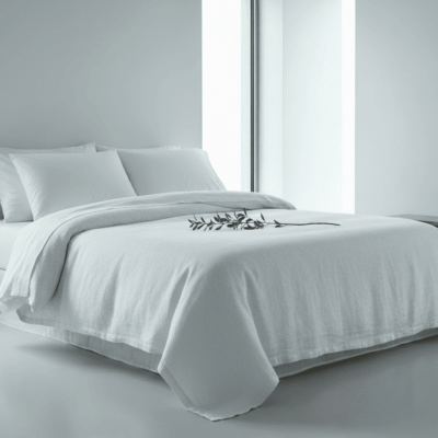 Ein modernes, minimalistisches Schlafzimmer mit Leinen Bettwäsche Fischgrat in Weiß von Vieböck 273, zwei Kissen, einem Ast mit grünen Blättern auf dem Bett, großen Fenstern und einer einfachen Holzbank vor einer schlichten weißen Wand.