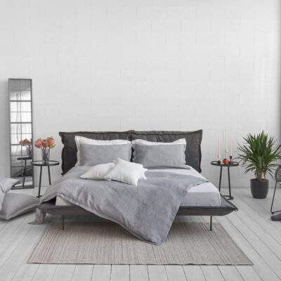 Modern bedroom with a grey bed in Arctica Classic Linen Bedding by Geniksa, white and grey bedding, pillows on the floor, a side table with candles and a plant, a chair and a tall mirror against a white wall and light wooden floor.
