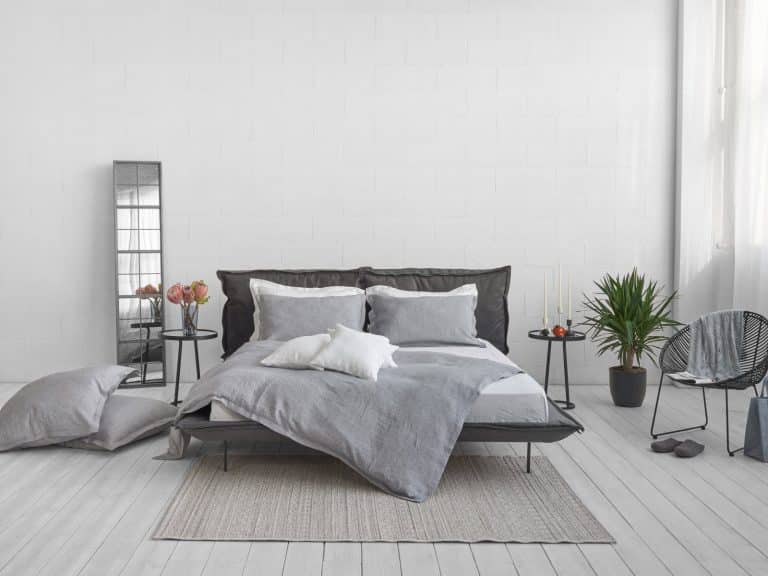 Modern bedroom with a grey bed in Arctica Classic Linen Bedding by Geniksa, white and grey bedding, pillows on the floor, a side table with candles and a plant, a chair and a tall mirror against a white wall and light wooden floor.