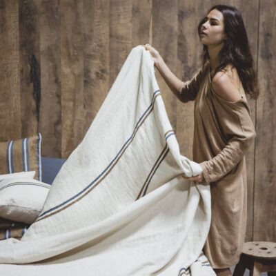 Eine Frau in einem lockeren, hellbraunen Kleid macht ein Bett mit cremefarbener und blau gestreifter Bettwäsche mit dem Auburn Leinen Coverlet und Kissen von Libeco in einem rustikalen Raum mit Holzwänden und gestreiften Kissen.