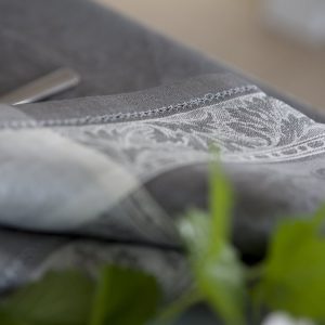 Nahaufnahme eines gefalteten grauen Stoffes mit einem verschlungenen weißen Blumenmuster, der auf einer Fläche mit grünen Blättern im Vordergrund ruht.