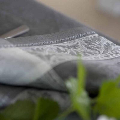 Nahaufnahme eines gefalteten grauen Stoffes mit einem verschlungenen weißen Blumenmuster, der auf einer Fläche mit grünen Blättern im Vordergrund ruht.