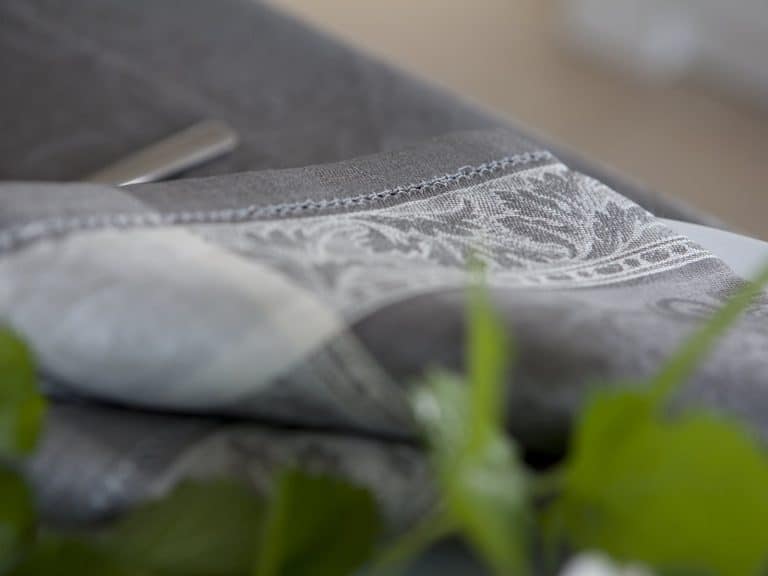 Close-up of a folded gray fabric with an intricate white floral pattern resting on a surface with green leaves in the foreground.