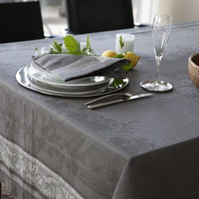 Auf einem Esstisch stehen die Tischdecke aus Leinen Damast Chrysantheme von Hoffmann, gestapelte weiße Teller, eine graue gefaltete Serviette, Silberbesteck, Glas, Kerze, ein geflochtener Korb und ein kleines Arrangement aus grünen Blättern mit Zitronen.