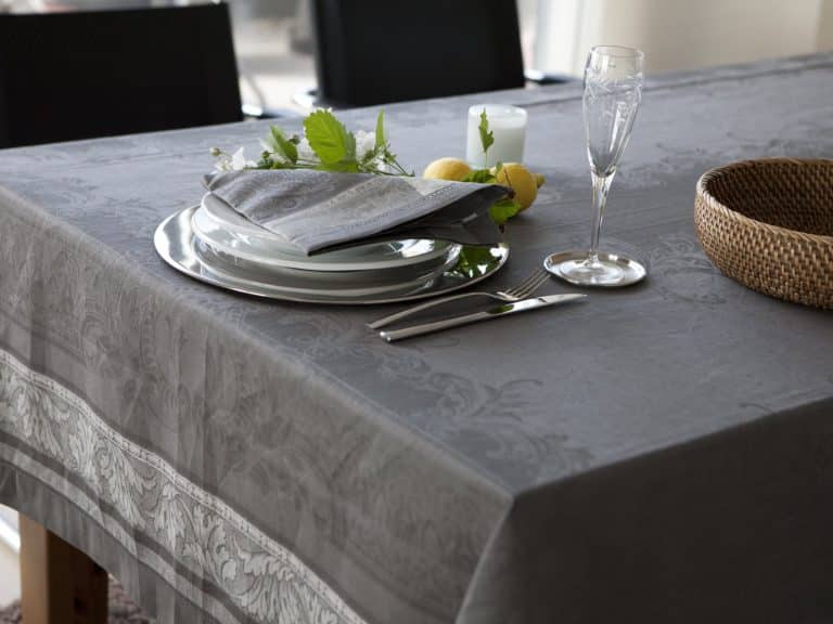 Auf einem Esstisch stehen die Tischdecke aus Leinen Damast Chrysantheme von Hoffmann, gestapelte weiße Teller, eine graue gefaltete Serviette, Silberbesteck, Glas, Kerze, ein geflochtener Korb und ein kleines Arrangement aus grünen Blättern mit Zitronen.