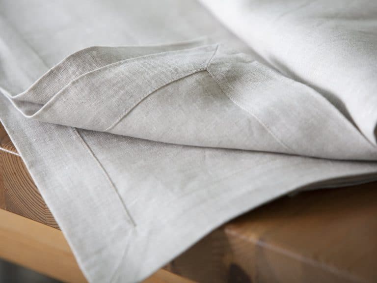 A close-up of a light gray linen tablecloth draped neatly over a wooden table, with neat seams and a textured fabric surface.