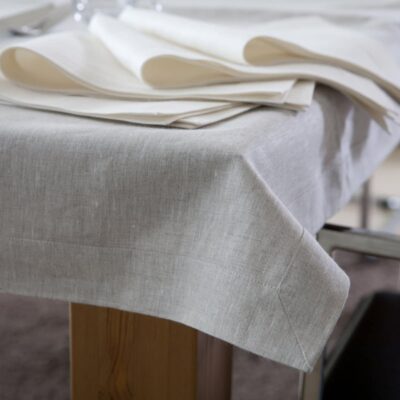 Nahaufnahme einer beigen Leinentischdecke, die einen Holztisch bedeckt, mit ordentlich gefalteten cremefarbenen Servietten, die am Rand platziert sind. Ein Metallstuhl ist teilweise neben dem Tisch zu sehen.