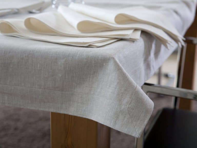 Close-up of a beige linen tablecloth covering a wooden table, with neatly folded cream-colored napkins placed around the edge. A metal chair is partially visible next to the table.