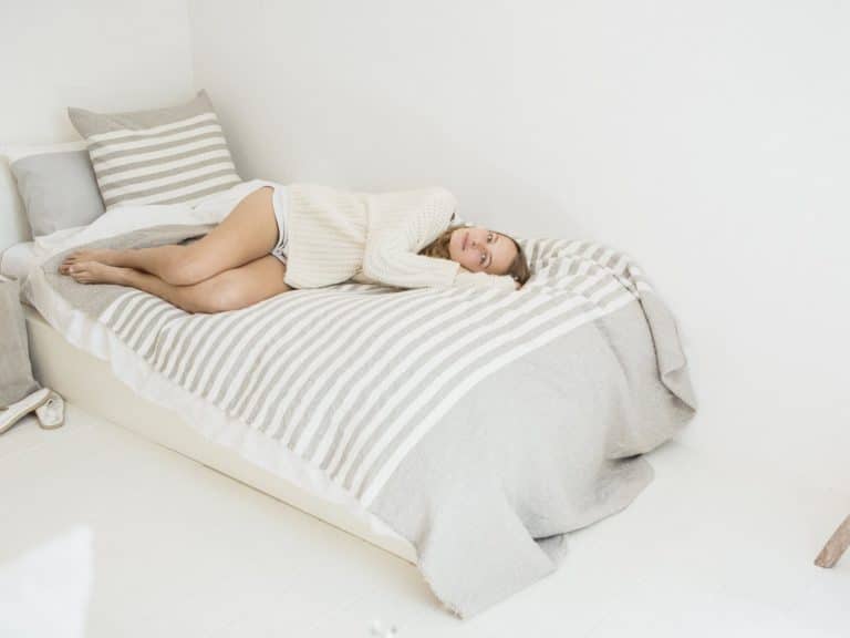 Eine Frau in einem cremefarbenen Pullover und Shorts entspannt sich auf einem gestreiften Bett in einem minimalistischen weißen Zimmer, das mit dem Camille Leinen Plaid von Libeco drapiert ist, und blickt nachdenklich in die Ferne.