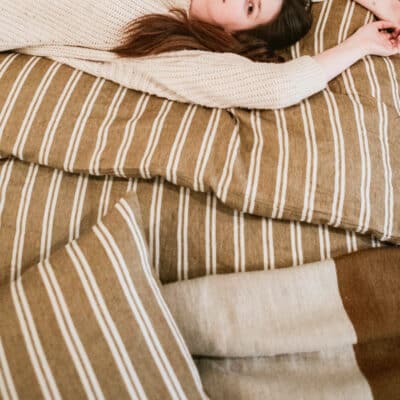 Eine Frau in einem cremefarbenen Pullover liegt entspannt auf einem Bett mit braunem und weißem Canal Stripe Leinen Bettwäsche von Libeco, die Arme über den Kopf gestreckt.