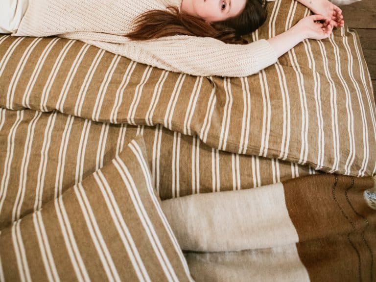 A woman in a cream-colored sweater lies relaxed on a bed with brown and white Canal Stripe linen bedding from Libeco, her arms stretched over her head.