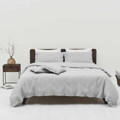 A neatly made bed with Caroline Buttons Linen Bedding by Geniksa in light grey and two pillows stands against a white wall. To the left is a dark wooden nightstand with a vase of dried branches, to the right a basket and a folded blanket.
