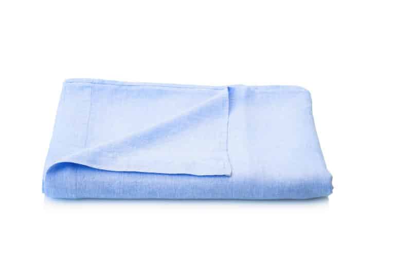 A folded Celeste linen bed sheet from Geniksa is shown on a white background, with light blue fabric and a partially folded corner.