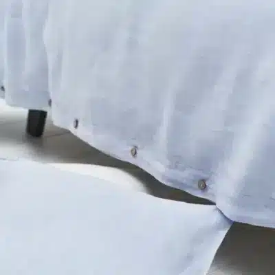 Close-up of a light blue Cygna Classic Linen Sheet by Geniksa with brown edge buttons, shown with a matching pillow and bedspread on a white floor.