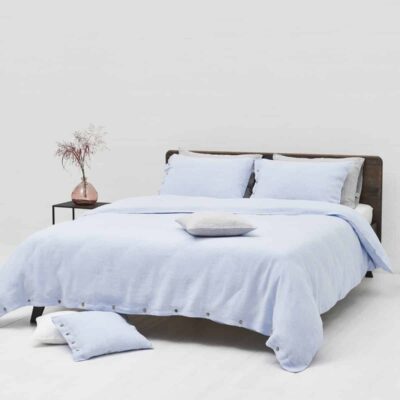 A neatly made bed with light blue Cygna Buttons Linen Bedding by Geniksa stands in a minimalist room, beside a small table with a pink vase of dried flowers, with two black decorative objects on the floor nearby.