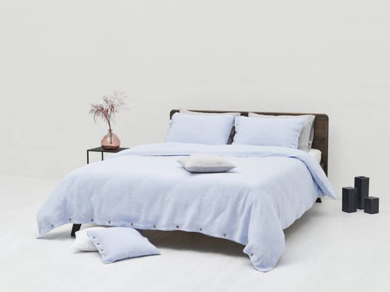 A neatly made bed with light blue Cygna Buttons Linen Bedding by Geniksa stands in a minimalist room, beside a small table with a pink vase of dried flowers, with two black decorative objects on the floor nearby.