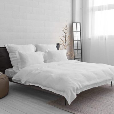 Minimalist bedroom with a low bed with white bedding and pillows, a brown stool with a blanket, a floor mirror, a bare decorative tree with lights and a large window with sheer curtains.