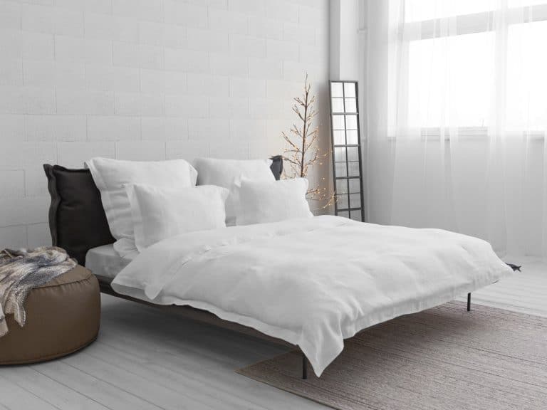 Minimalist bedroom with a low bed with white bedding and pillows, a brown stool with a blanket, a floor mirror, a bare decorative tree with lights and a large window with sheer curtains.