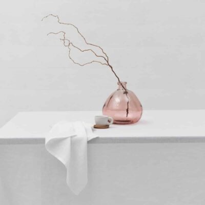 A minimalist scene with the Cygna linen tablecloth by Geniksa, a pink glass vase with bare branches, a white cup and saucer and a draped napkin, all against a plain white background.
