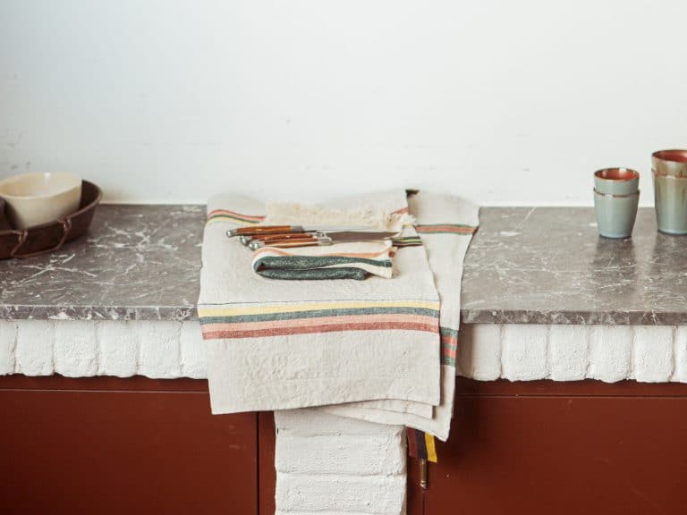 Ein gefaltetes gestreiftes Tuch mit Messern und einem Gemüseschäler liegt auf einer Marmorarbeitsplatte. Zwei Keramiktassen und eine Schüssel stehen daneben an einer schlichten weißen Wand.