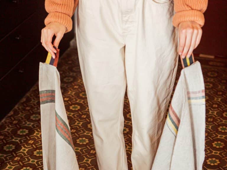 Eine Person in einem pfirsichfarbenen Pullover und einer cremefarbenen Hose hält zwei bunt gestreifte, wiederverwendbare Taschen - eine mit dem Design Dock Green Stripe - reines Leinen für die Küche von Libeco. Darunter ist ein gemusterter Fliesenboden zu sehen.
