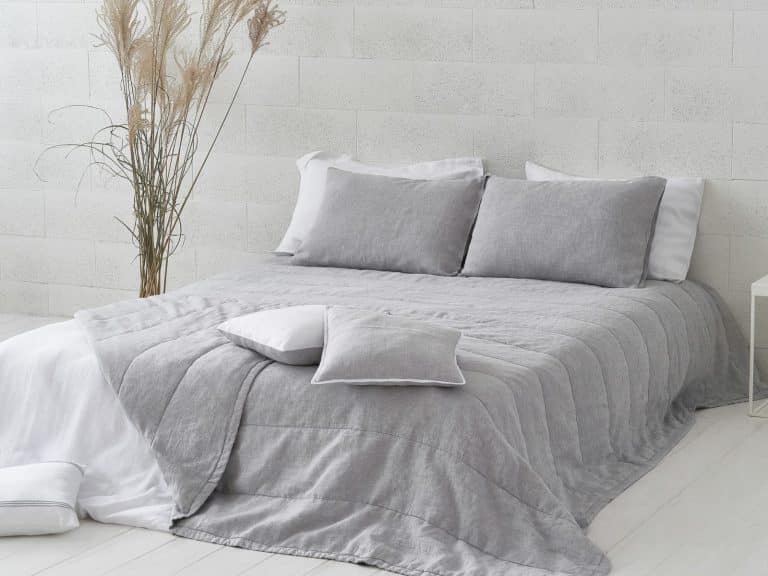 A neatly made bed with light gray bedding and pillows presents the Caroline linen bedspread from Geniksa against a white textured wall. A vase of tall dried grass adds calm and minimalist style to the bedroom.