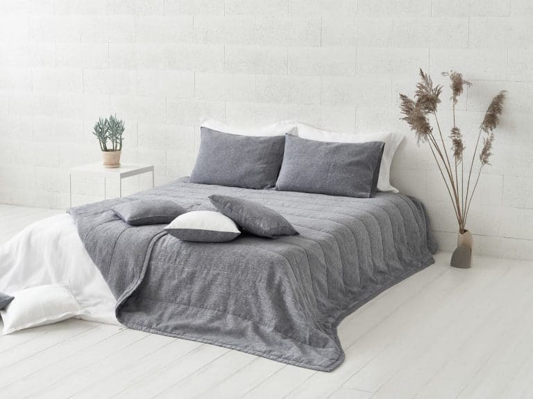 Ein minimalistisches Zimmer mit einem ordentlich gemachten Bett mit grauer Heron Classic Leinen Tagesdecke von Geniksa, weißen Akzenten, einer weißen Backsteinwand, einem kleinen Beistelltisch mit einer Topfpflanze und hohen getrockneten Gräsern in einer Vase.