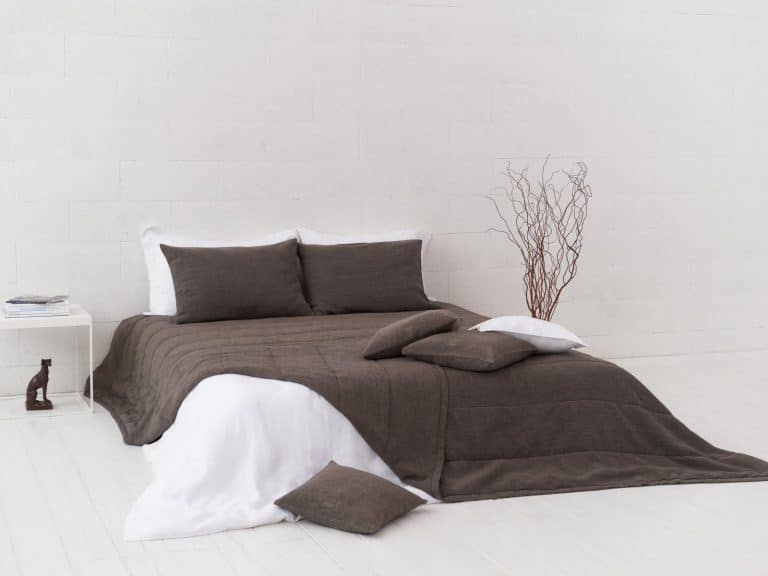 Ein ordentlich gemachtes Bett mit dunkelgrauer Bettwäsche, darunter die Jungle Classic Leinen Tagesdecke von Geniksa, weiße Laken, drei passende Kissen auf dem Bett und zwei auf dem Boden. Auf einem weißen Beistelltisch stehen eine kleine Skulptur und Bücher in einem minimalistischen weißen Raum.