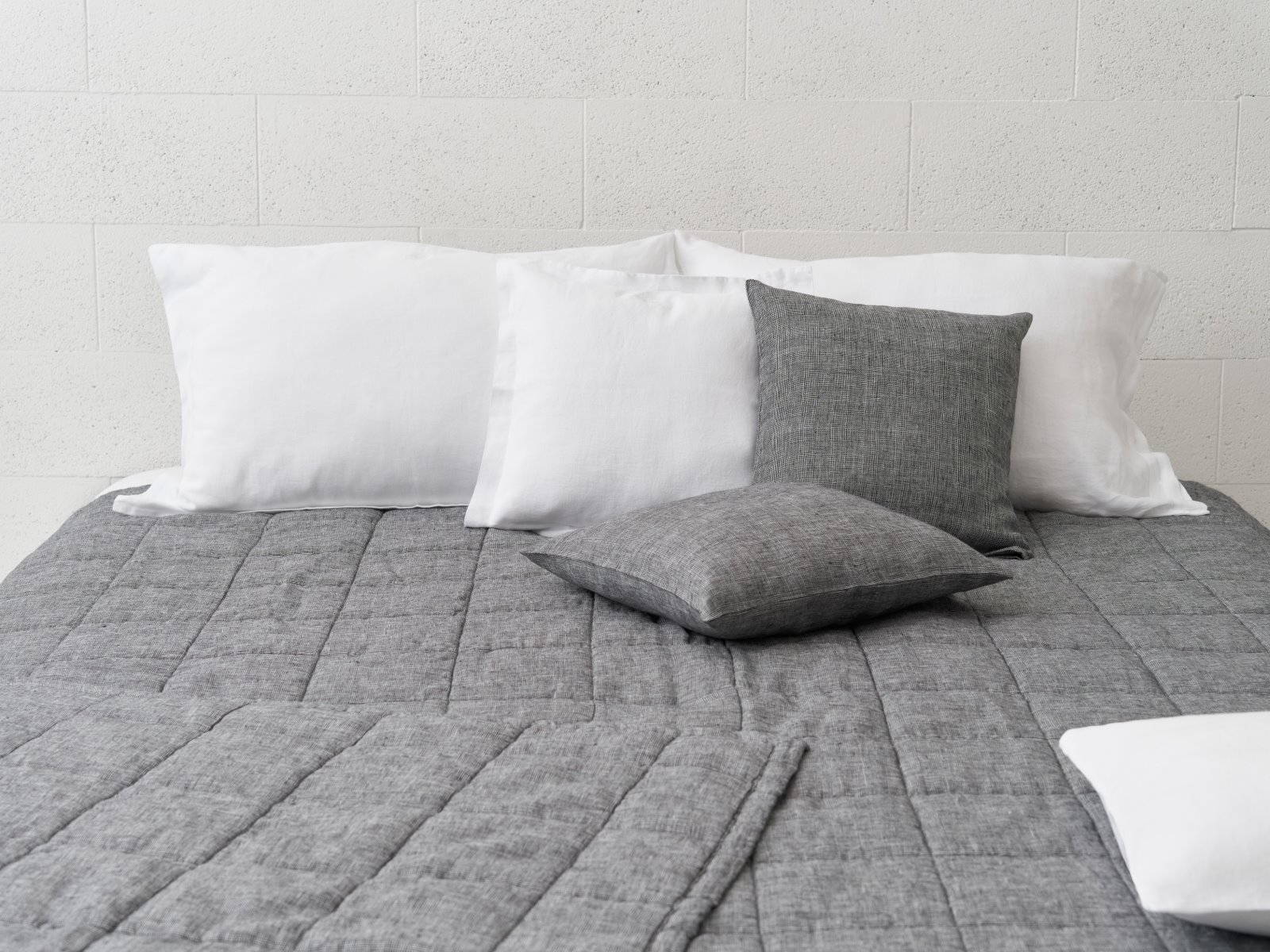 Ein ordentlich gemachtes Bett mit der Scotia Leinen Tagesdecke von Geniksa, zwei grauen Zierkissen und drei großen weißen Kissen vor einer weißen, strukturierten Wand.