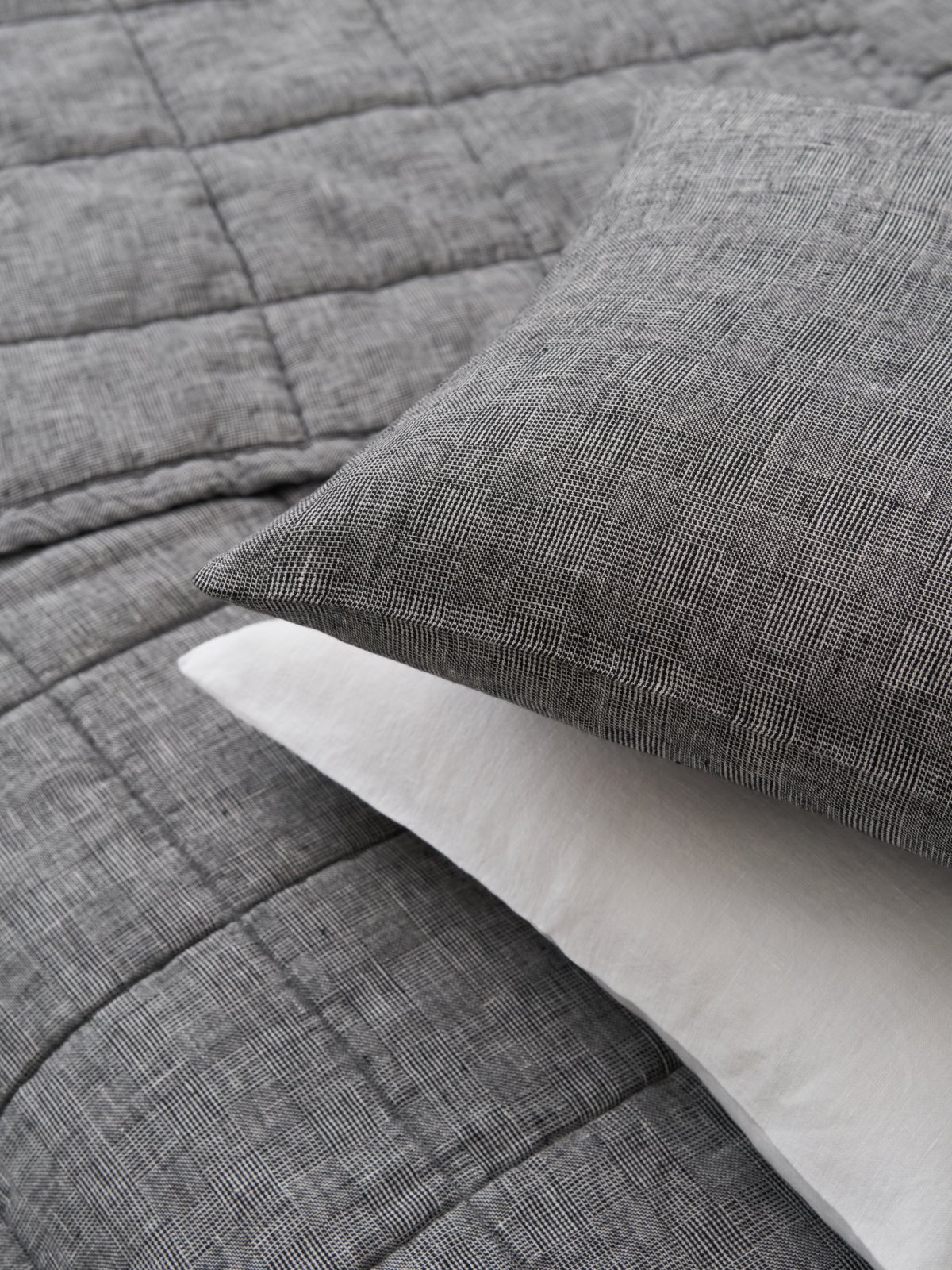 Die Nahaufnahme eines ordentlich gemachten Bettes zeigt die Scotia Leinen Tagesdecke von Geniksa, eine dunkelgraue, strukturierte Bettdecke mit einem dezenten Gittermuster. Ihr modernes, minimalistisches Design unterstreicht den zeitgenössischen Stil des Zimmers.