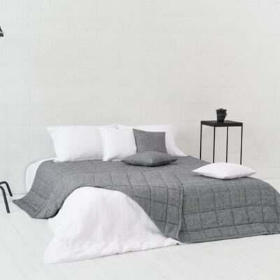 Ein minimalistisches Schlafzimmer mit einem niedrigen Bett in Grau und Weiß, mit der Scotia Leinen Tagesdecke von Geniksa, drei Kissen, einem schwarzen Beistelltisch mit einem kleinen Gegenstand und einer Studiolampe auf einem Ständer vor weißen Wänden und Böden.