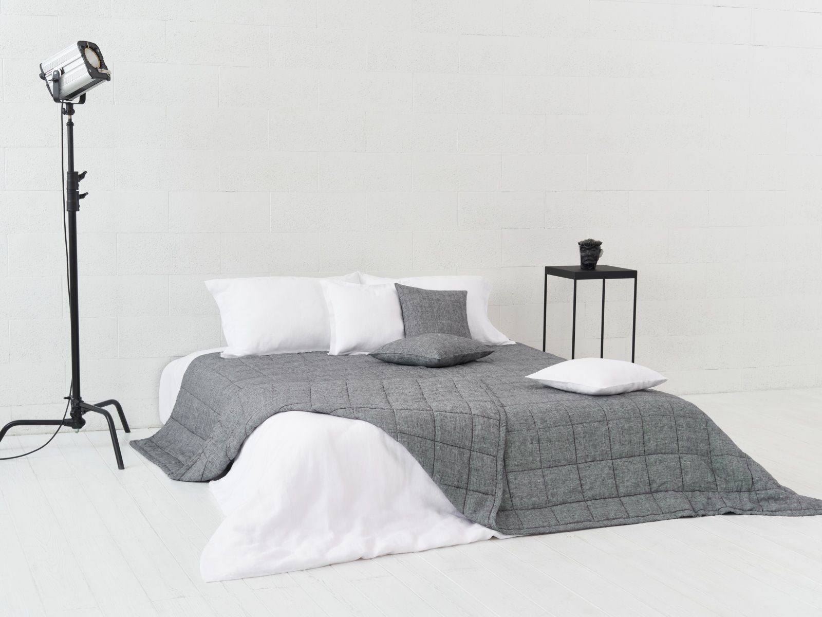 Ein minimalistisches Schlafzimmer mit einem niedrigen Bett in Grau und Weiß, mit der Scotia Leinen Tagesdecke von Geniksa, drei Kissen, einem schwarzen Beistelltisch mit einem kleinen Gegenstand und einer Studiolampe auf einem Ständer vor weißen Wänden und Böden.