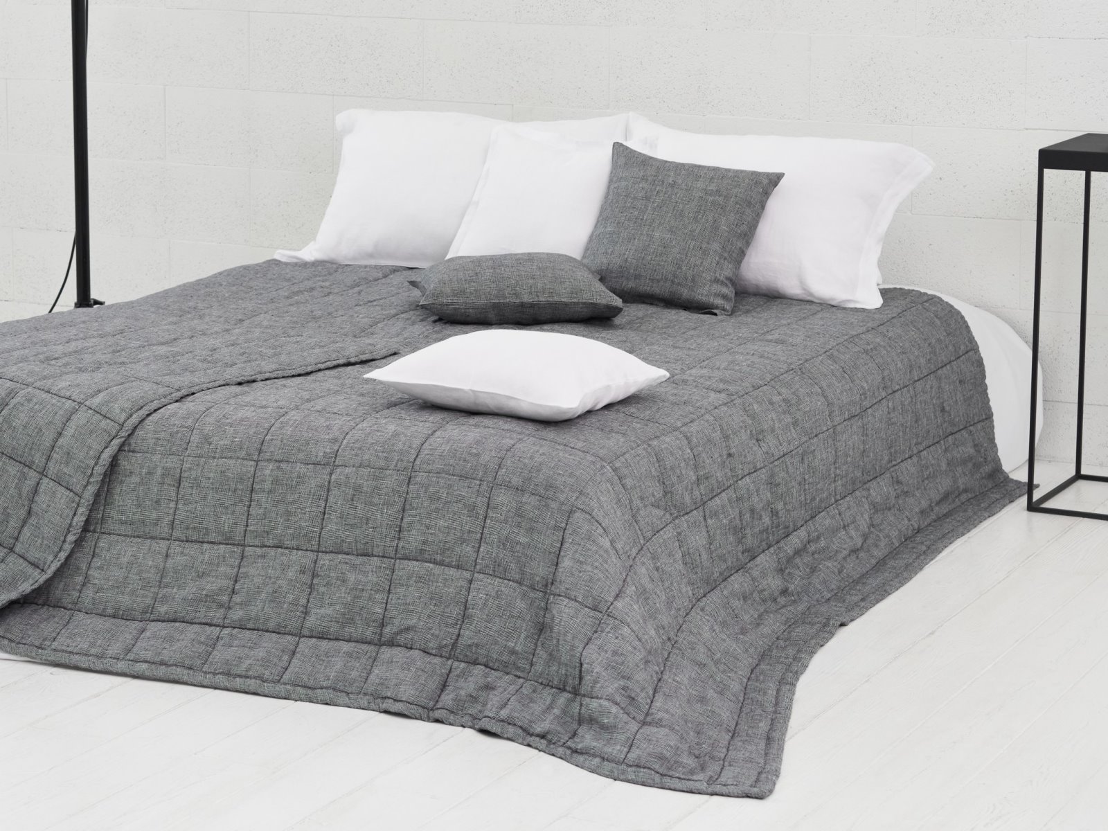 Ein ordentlich gemachtes Bett mit der Scotia Leinen Tagesdecke von Geniksa in Grau, zwei weißen Kissen, einem grauen Kissen und einem weiteren weißen Kissen darauf. Das Bett steht vor einer weißen Wand mit einer schwarzen Lampe und einem Nachttisch daneben.