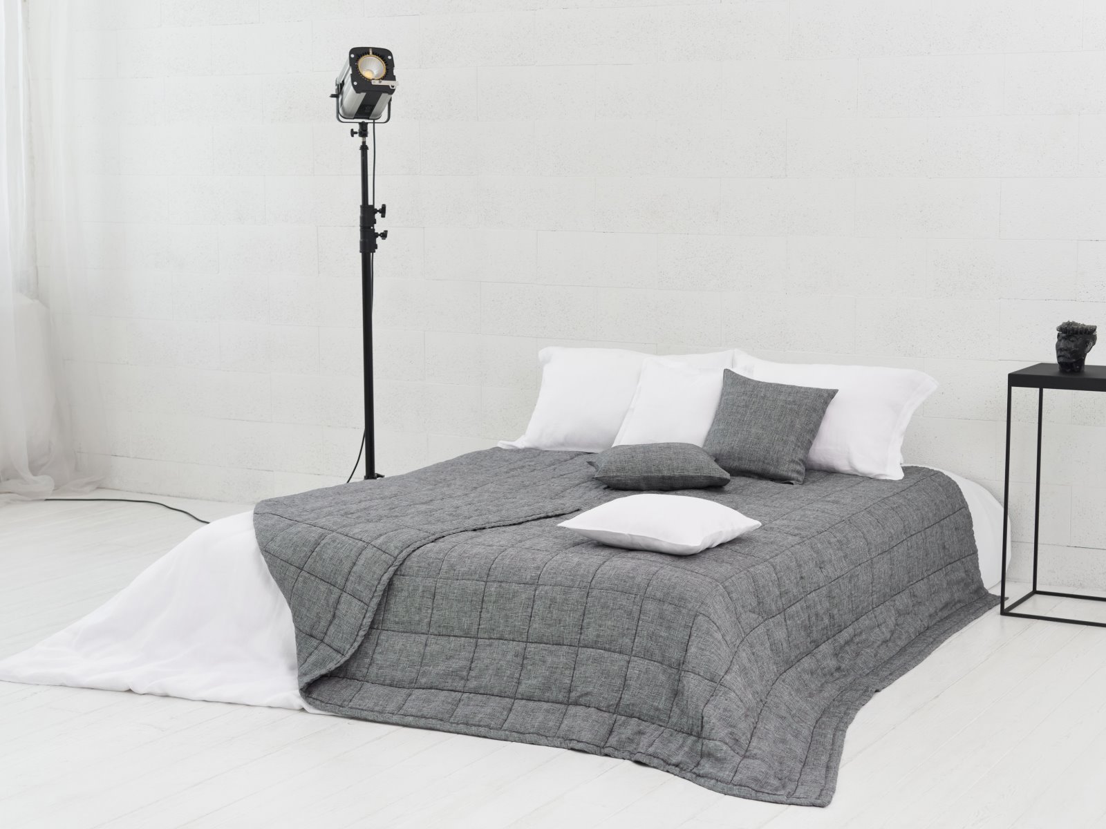 Ein modernes, minimalistisches Schlafzimmer mit einem niedrigen Bett, das von der Scotia Leinen Tagesdecke von Geniksa gekrönt wird, weißen und grauen Kissen, einem schwarzen Beistelltisch und einer hohen Studioleuchte vor einer weißen Backsteinwand.