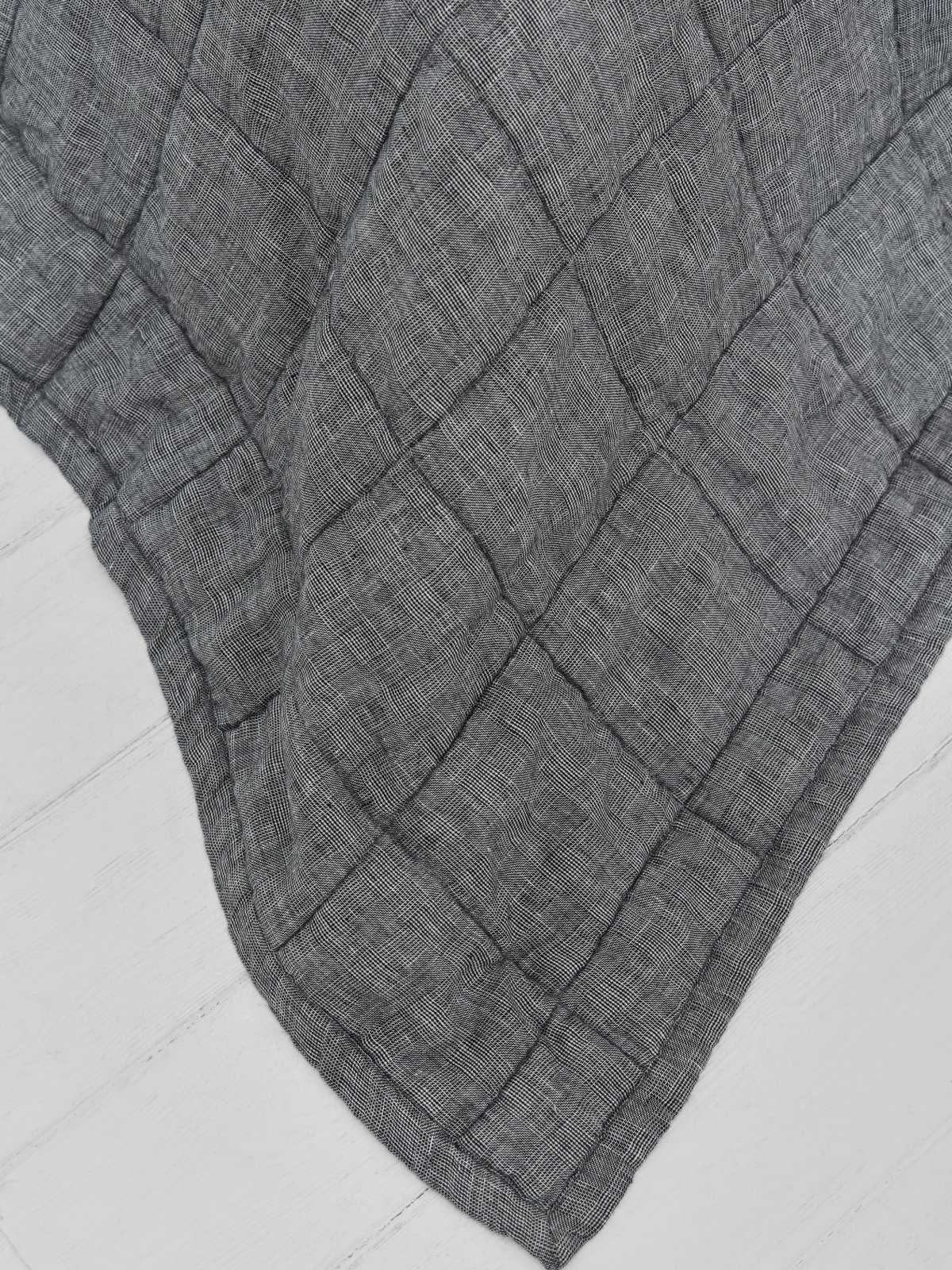 Eine Nahaufnahme der Scotia Leinen Tagesdecke von Geniksa in Dunkelgrau, die ihr gestepptes Gittermuster auf einer hellen Holzoberfläche zur Geltung bringt.