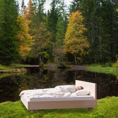 A person sleeps outdoors beside a tranquil forest pond on a bed with the Pure Bamboo Summer Duvet by HEFEL, surrounded by lush greenery and autumn-coloured trees.