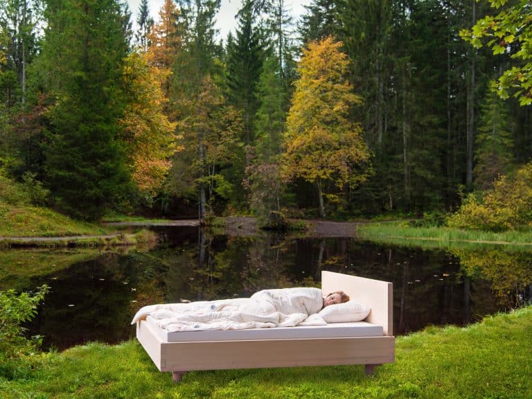 One person sleeps outdoors next to a tranquil forest pond on a bed with the Pure Bamboo summer blanket from HEFEL, surrounded by lush greenery and autumn-colored trees.